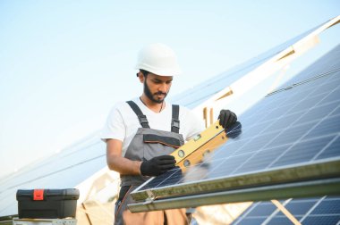 Kasklı ve kahverengi tulumlu erkek Arap mühendis güneş panellerindeki direnci kontrol ediyor. İstasyonda çalışan Hintli adam.