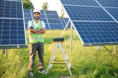 Kasklı ve kahverengi tulumlu erkek Arap mühendis güneş panellerindeki direnci kontrol ediyor. İstasyonda çalışan Hintli adam.