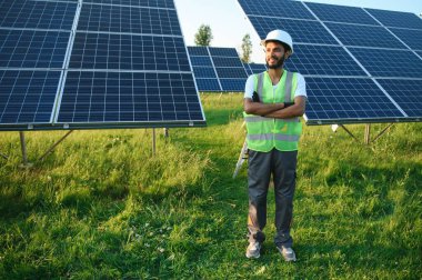 Kasklı ve kahverengi tulumlu erkek Arap mühendis güneş panellerindeki direnci kontrol ediyor. İstasyonda çalışan Hintli adam.