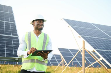 Beyaz gömlekli ve şapkalı Afro-Amerikalı bir mühendis güneş panelleri üzerinde çalışıyor. Güneş enerjisi yenilenebilir enerji kavramı