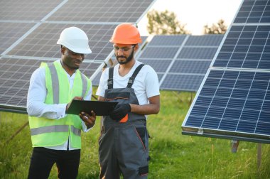 Güvenlik kasklı çok ırklı bir grup insan güneş istasyonunda duruyor ve ellerinde büyük çizimler olan kameralara bakıyor. İki mühendis ve teknisyen dışarıdaki panelleri inceliyor..