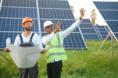 Güvenlik kasklı çok ırklı bir grup insan güneş istasyonunda duruyor ve ellerinde büyük çizimler olan kameralara bakıyor. İki mühendis ve teknisyen dışarıdaki panelleri inceliyor..