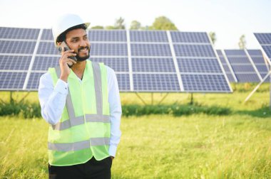 Yeşil yelek ve kask giyen genç erkek teknisyen, sahada duran güneş panellerinin bakımı ve kurulumu hakkında telefonda konuşuyor..