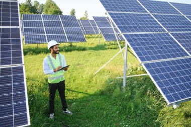 Güneş panellerinin yanında duran tabletli genç erkek mühendis, temiz mavi gökyüzü geçmişe sahip tarım arazisi, yenilenebilir enerji, temiz enerji