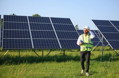 Mavi gökyüzüne karşı güneş panelleri yakınında duran beyaz başlıklı genç Hintli teknisyen portresi. Endüstriyel işçi güneş sistemi kurulumu, yenilenebilir yeşil enerji üretimi konsepti.