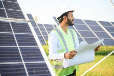 Mavi gökyüzüne karşı güneş panelleri yakınında duran beyaz başlıklı genç Hintli teknisyen portresi. Endüstriyel işçi güneş sistemi kurulumu, yenilenebilir yeşil enerji üretimi konsepti.
