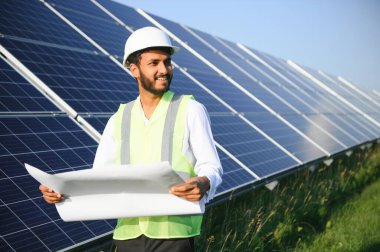 Genç Hintli teknisyen ya da yönetici güneş panelinin yanında duran resmi giysiler giyiyor. Yenilenebilir enerji, çapraz duran adam, boşluğu kopyala.