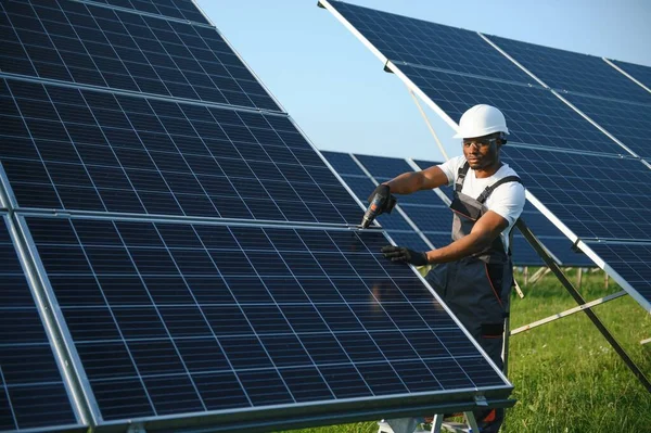 Competent energy engineer in grey overalls and orange helmet checking solar panels while walking on field. African american man carrying clipboard and container with instruments.