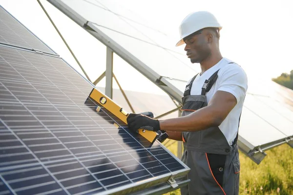 stock image Competent energy engineer in grey overalls and orange helmet checking solar panels while walking on field. African american man carrying clipboard and container with instruments.