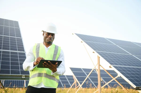 An African-American engineer in a white shirt and hard hat is working on a field of solar panels. Solar renewable energy concept