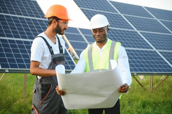 Group of multi ethnic people in safety helmets standing at solar station and looking on camera holding big drawings. Two engineers and technician examining plan of panels outdoors.