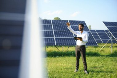 Güneş panellerinin yanında duran tabletli genç erkek mühendis, temiz mavi gökyüzü geçmişe sahip tarım arazisi, yenilenebilir enerji, temiz enerji