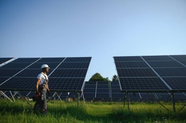 Gri tulumlu ehil enerji mühendisi ve beyaz miğfer sahada yürürken güneş panellerini kontrol ediyor. Elinde enstrümanlarla pano ve konteynır taşıyan Afrikalı Amerikalı bir adam..