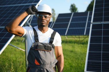 Beyaz kasklı ve gri tulumlu Afro-Amerikan adam güneş panellerinin arasında duruyor..