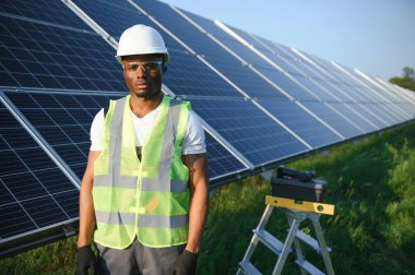 Beyaz kasklı ve gri tulumlu Afro-Amerikan adam güneş panellerinin arasında duruyor..