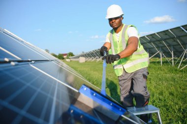 Gri tulumlu ve turuncu başlıklı ehil enerji mühendisi sahada yürürken güneş panellerini kontrol ediyor. Elinde enstrümanlarla pano ve konteynır taşıyan Afrikalı Amerikalı bir adam..