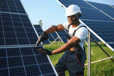 Güvenlik kaskı ve gözlüklü Afrikalı Amerikalı adam güneş panellerini tornavidayla sıkıyor. İstasyonda servis çalışması yaparken araçları kullanan yetkili teknisyen.