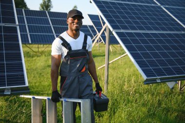 Koruyucu miğfer takmış yakışıklı Afrikalı Amerikalı zanaatkârın portresi. Üniformalı ve aletleri güneş panellerinin arasında duran bir adam..