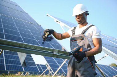 Koruyucu miğfer takmış yakışıklı Afrikalı Amerikalı zanaatkârın portresi. Üniformalı ve aletleri güneş panellerinin arasında duran bir adam..