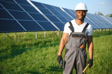 Beyaz kasklı ve gri tulumlu Afro-Amerikan adam güneş panellerinin arasında duruyor..