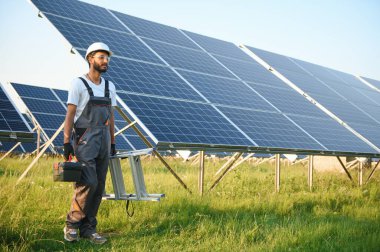 Kasklı ve kahverengi tulumlu erkek Arap mühendis güneş panellerindeki direnci kontrol ediyor. İstasyonda çalışan Hintli adam.