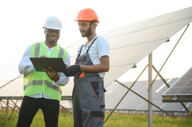 Güvenlik kasklı çok ırklı bir grup insan güneş istasyonunda duruyor ve ellerinde büyük çizimler olan kameralara bakıyor. İki mühendis ve teknisyen dışarıdaki panelleri inceliyor..