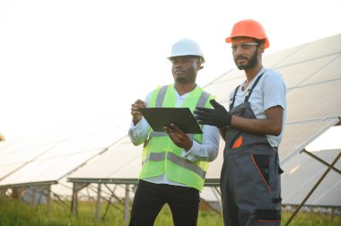 Güvenlik kasklı çok ırklı bir grup insan güneş istasyonunda duruyor ve ellerinde büyük çizimler olan kameralara bakıyor. İki mühendis ve teknisyen dışarıdaki panelleri inceliyor..