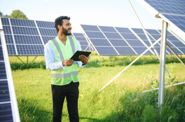 Güneş panellerinin yanında duran tabletli genç erkek mühendis, temiz mavi gökyüzü geçmişe sahip tarım arazisi, yenilenebilir enerji, temiz enerji
