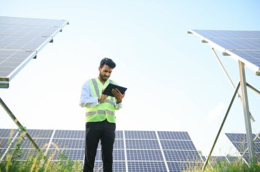 Genç Hintli erkek mühendisin portresi güneş panellerinin yanında duruyor, açık mavi gökyüzü arka planı, yenilenebilir ve temiz enerjisi var. beceri Hindistan, kopyalama alanı