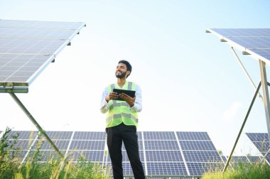 Genç Hintli teknisyen ya da yönetici güneş panelinin yanında duran resmi giysiler giyiyor. Yenilenebilir enerji, çapraz duran adam, boşluğu kopyala.