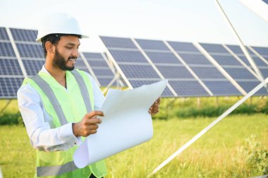 Mavi gökyüzüne karşı güneş panelleri yakınında duran beyaz başlıklı genç Hintli teknisyen portresi. Endüstriyel işçi güneş sistemi kurulumu, yenilenebilir yeşil enerji üretimi konsepti.