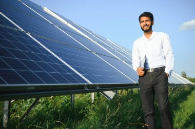 Genç Hintli teknisyen ya da yönetici güneş panelinin yanında duran resmi giysiler giyiyor. Yenilenebilir enerji, çapraz duran adam, boşluğu kopyala.