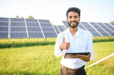 Genç Hintli teknisyen ya da yönetici güneş panelinin yanında duran resmi giysiler giyiyor. Yenilenebilir enerji, çapraz duran adam, boşluğu kopyala.