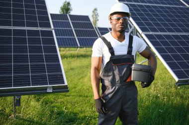 Gri tulumlu ve turuncu başlıklı ehil enerji mühendisi sahada yürürken güneş panellerini kontrol ediyor. Elinde enstrümanlarla pano ve konteynır taşıyan Afrikalı Amerikalı bir adam..