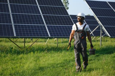 Koruyucu miğfer takmış yakışıklı Afrikalı Amerikalı zanaatkârın portresi. Üniformalı ve aletleri güneş panellerinin arasında duran bir adam..