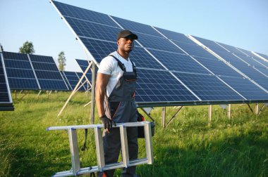 Beyaz kasklı ve gri tulumlu Afro-Amerikan adam güneş panellerinin arasında duruyor..