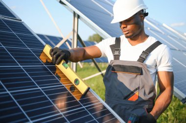 Gri tulumlu ve turuncu başlıklı ehil enerji mühendisi sahada yürürken güneş panellerini kontrol ediyor. Elinde enstrümanlarla pano ve konteynır taşıyan Afrikalı Amerikalı bir adam..