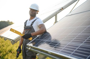 Koruyucu miğfer takmış yakışıklı Afrikalı Amerikalı zanaatkârın portresi. Üniformalı ve aletleri güneş panellerinin arasında duran bir adam..