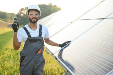 Kasklı ve kahverengi tulumlu erkek Arap mühendis güneş panellerindeki direnci kontrol ediyor. İstasyonda çalışan Hintli adam.