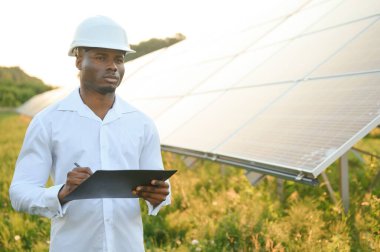 Beyaz gömlekli ve şapkalı Afro-Amerikalı bir mühendis güneş panelleri üzerinde çalışıyor. Güneş enerjisi yenilenebilir enerji kavramı