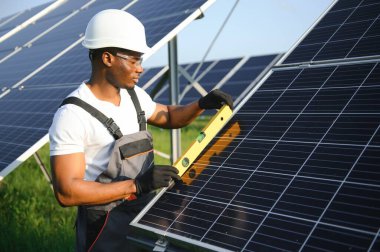 Beyaz kasklı ve gri tulumlu Afro-Amerikan adam güneş panellerinin arasında duruyor..