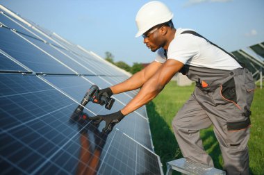 Güvenlik kaskı ve gözlüklü Afrikalı Amerikalı adam güneş panellerini tornavidayla sıkıyor. İstasyonda servis çalışması yaparken araçları kullanan yetkili teknisyen.