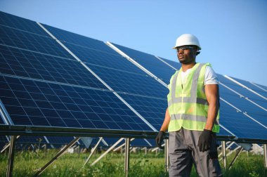 Beyaz kasklı ve gri tulumlu Afro-Amerikan adam güneş panellerinin arasında duruyor..