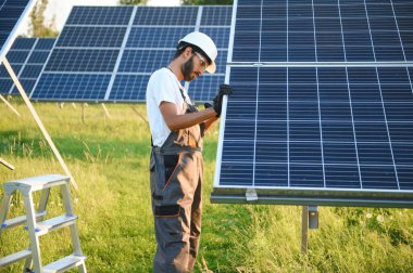 Kasklı ve kahverengi tulumlu erkek Arap mühendis güneş panellerindeki direnci kontrol ediyor. İstasyonda çalışan Hintli adam.