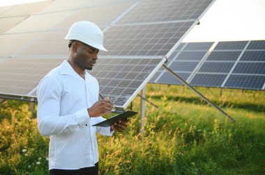 Beyaz gömlekli ve şapkalı Afro-Amerikalı bir mühendis güneş panelleri üzerinde çalışıyor. Güneş enerjisi yenilenebilir enerji kavramı