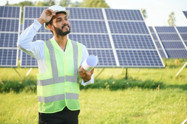 Portrait of Young indian man technician wearing white hard hat standing near solar panels against blue sky.Industrial worker solar system installation, renewable green energy generation concept