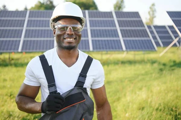 Gri tulumlu ehil enerji mühendisi ve beyaz miğfer sahada yürürken güneş panellerini kontrol ediyor. Elinde enstrümanlarla pano ve konteynır taşıyan Afrikalı Amerikalı bir adam..