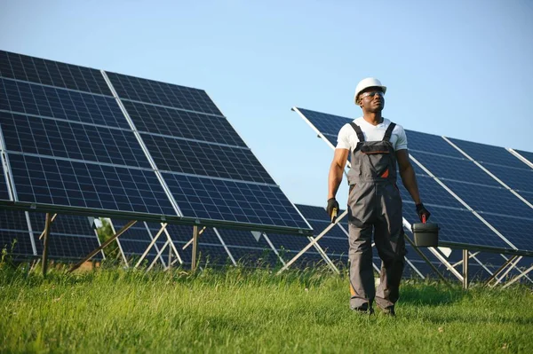 Gri tulumlu ehil enerji mühendisi ve beyaz miğfer sahada yürürken güneş panellerini kontrol ediyor. Elinde enstrümanlarla pano ve konteynır taşıyan Afrikalı Amerikalı bir adam..