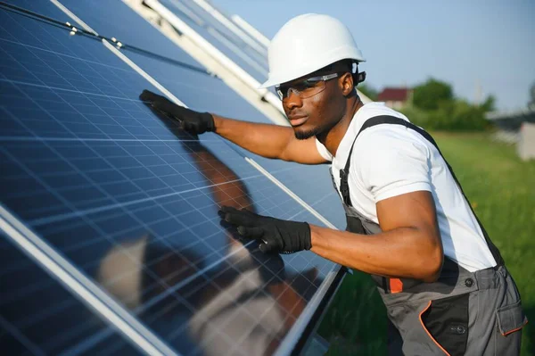 Gri tulumlu ve turuncu başlıklı ehil enerji mühendisi sahada yürürken güneş panellerini kontrol ediyor. Elinde enstrümanlarla pano ve konteynır taşıyan Afrikalı Amerikalı bir adam..