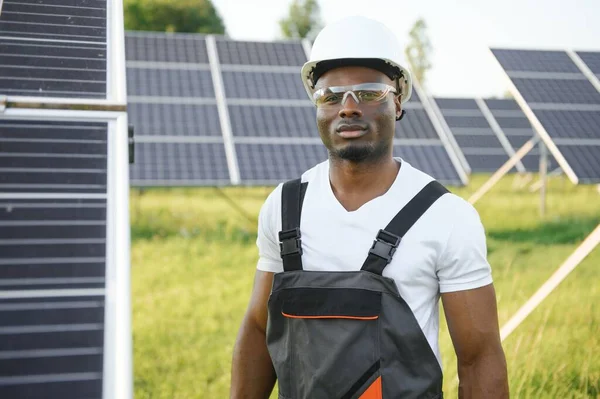 Beyaz kasklı ve gri tulumlu Afro-Amerikan adam güneş panellerinin arasında duruyor..